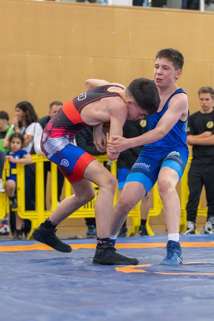 Two teenage wrestlers grapple intensely on a blue mat, one in black-red, the other in blue, spectators watching behind barriers.
