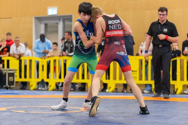 Two young wrestlers in a tense clinch on a blue mat, one wearing a NED singlet, referee watching nearby.