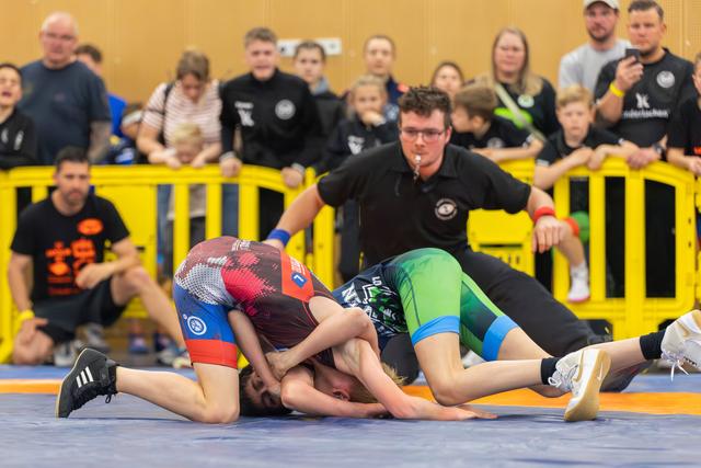 Two young wrestlers grapple intensely on a blue mat, watched closely by a referee and a crowd of spectators.