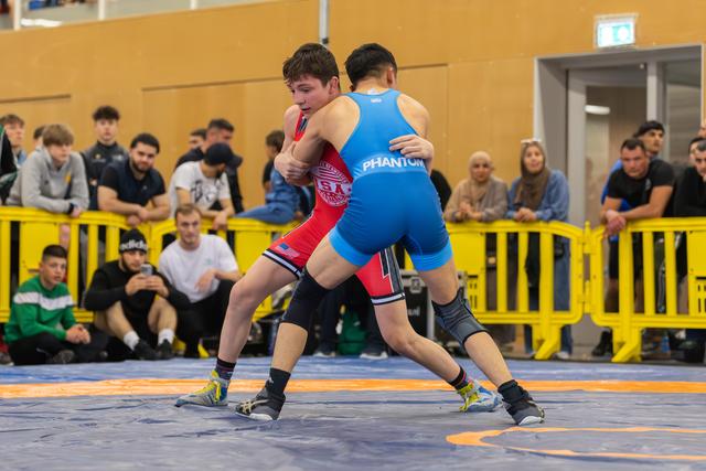 Two young wrestlers lock up intensely on a blue mat, one in red USA singlet, one in blue Phantom, crowd watching.