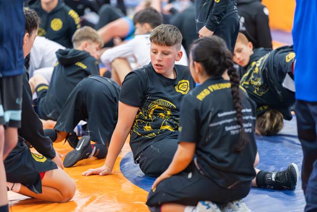 Young wrestlers in black and gold team shirts sit on the mat, chatting between matches at an indoor sports event.