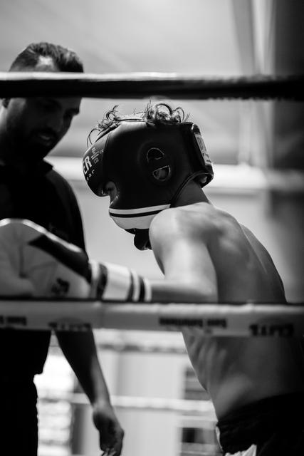 Boxer wearing protective headgear stands in the ring during a match, coach visible in background through the ropes