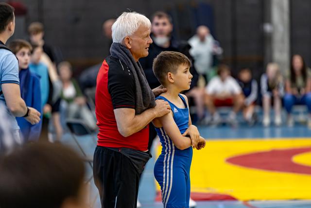 Older coach with white hair comforts young wrestler in blue singlet on yellow mat during indoor tournament