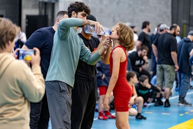 Coach pours water into mouth of young wrestler in red singlet during break, with spectators and athletes in background