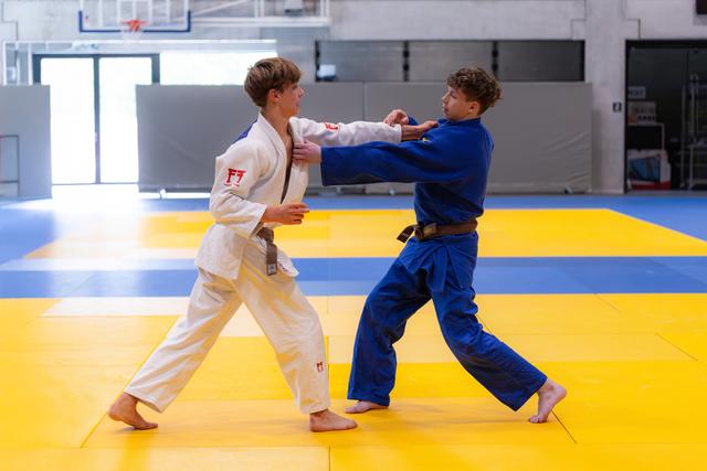 Two young judoka practice gripping techniques on yellow training mats in a modern sports facility, wearing white and blue gi
