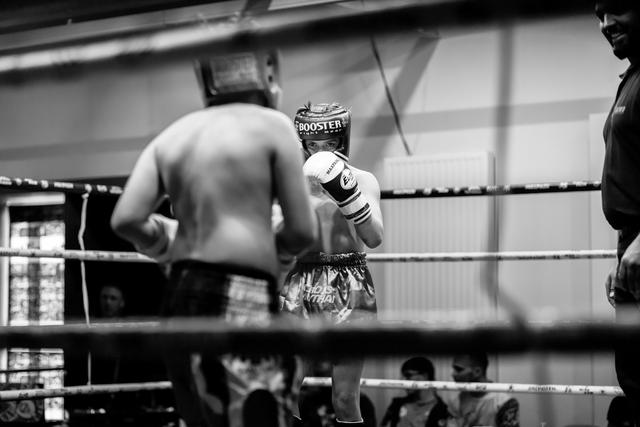 Muay Thai fighter with Booster headgear stands in ring facing shirtless opponent, coach observing from corner