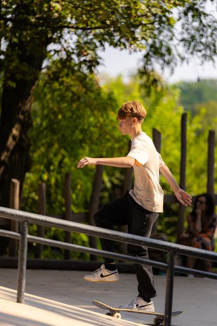 Young skater balances on rail during grind trick at outdoor skatepark, focused expression, surrounded by lush greenery