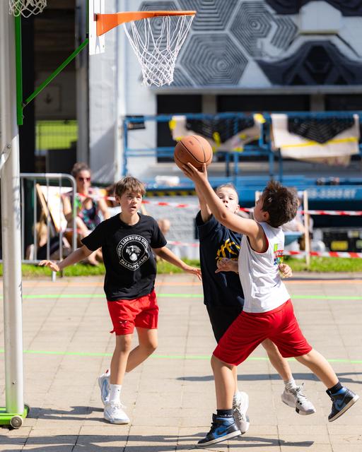 Three young players compete in an outdoor basketball game, one boy shooting while others defend on a sunny court