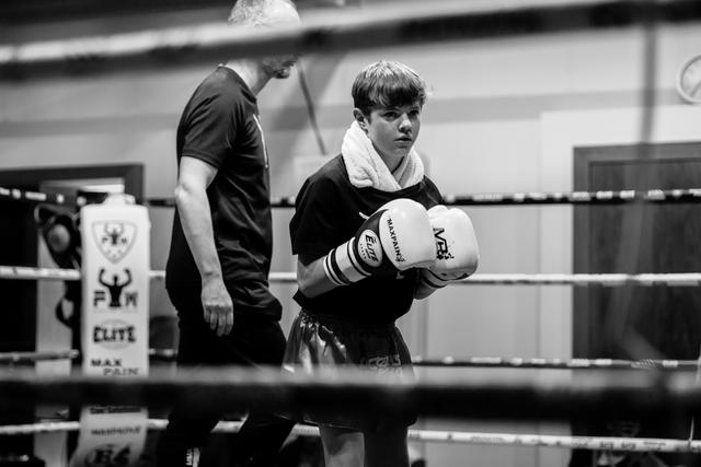 Young boxer with white towel around neck stands focused in ring wearing boxing gloves, coach visible in background