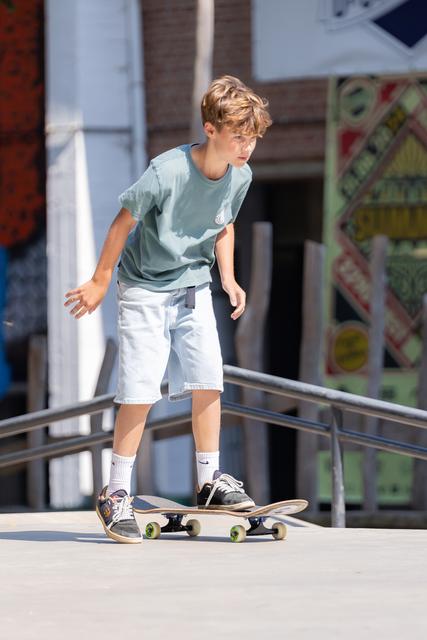 Young boy balances on skateboard with focused concentration, wearing light blue shirt and white shorts at outdoor skate park