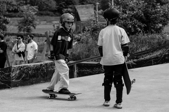 Young skateboarder in helmet rides past watching peer at outdoor skate park, surrounded by spectators and greenery
