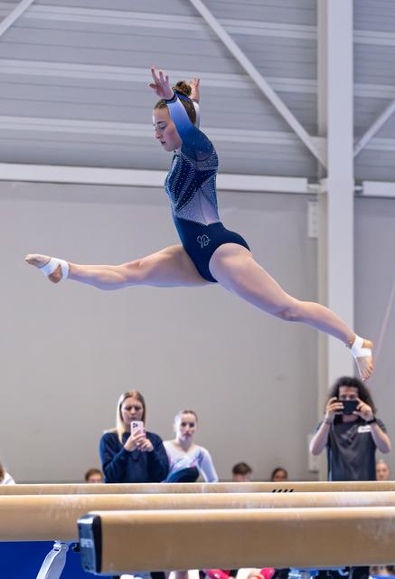 Gymnast performs a split leap above the balance beam while spectators watch and photograph from below
