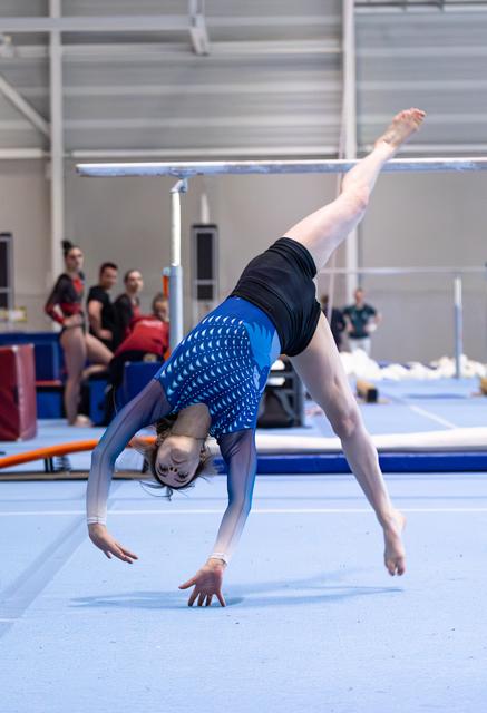 Female athlete executing a dramatic backbend on the floor mat while spectators watch from the sidelines during a meet