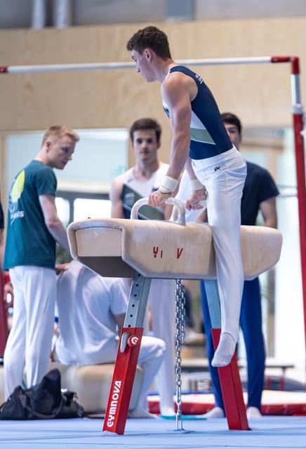 Male athlete gripping pommel horse handles, preparing for routine while coach observes and teammates watch in background