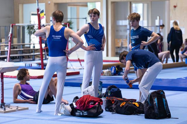 Young male athletes in blue uniforms gather on training floor, some standing while others rest near their bags during practice