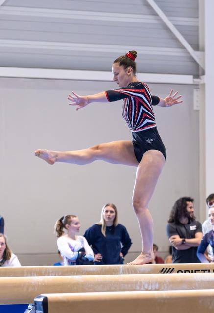 Gymnast performs a leap on balance beam with arms extended, spectators watching in background at indoor facility