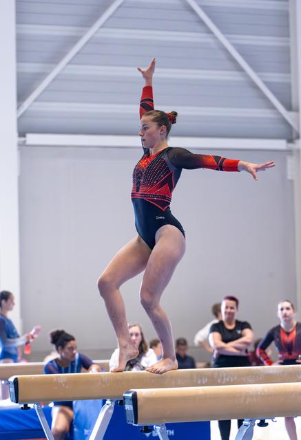 Female gymnast performing on balance beam with one arm raised and one extended, wearing navy and red leotard with rhinestones