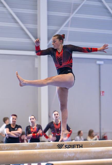 Athlete performing on balance beam with extended leg and arms, teammates in matching outfits watching in background