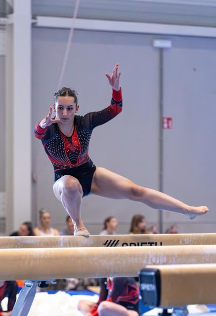 Gymnast performs focused routine on balance beam with extended leg and raised arms, displaying concentration and control