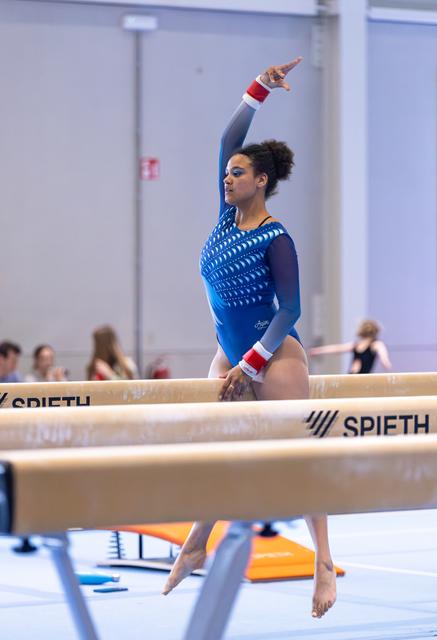 Female gymnast performing on balance beam with one arm raised, wearing blue leotard with red and white accents