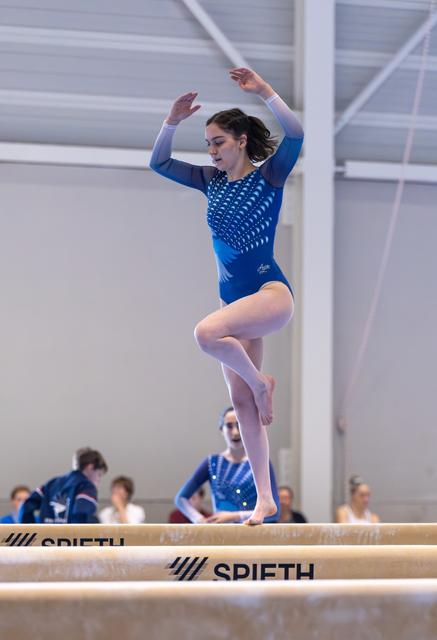 Athlete performs precise leap on balance beam in blue sparkled leotard, arms gracefully raised, showing intense concentration