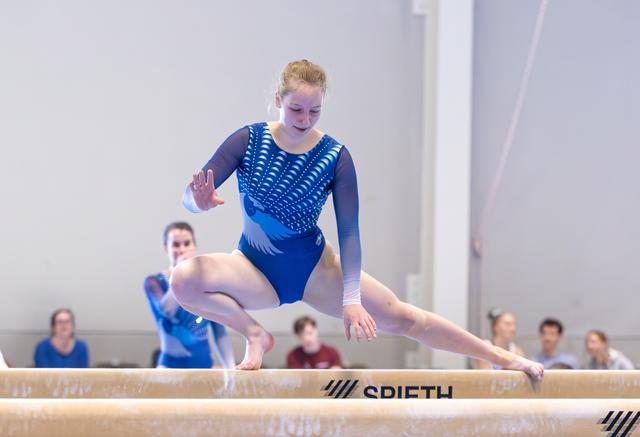 Young athlete performs a split leap on balance beam with focused expression, teammates visible in background