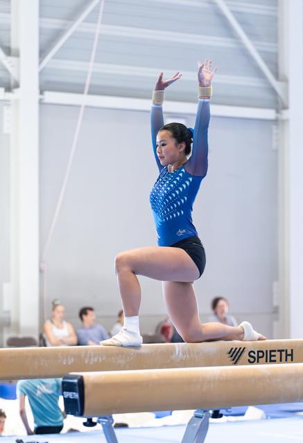 Gymnast performing on balance beam with arms raised overhead, wearing blue leotard, in focused athletic stance