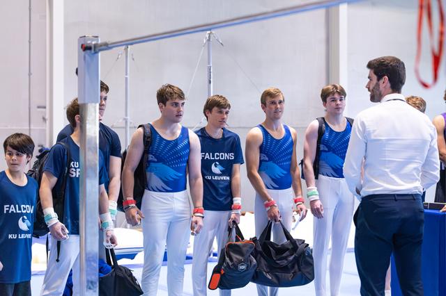 Coach in white shirt instructs four young male athletes in blue uniforms holding gym bags before their performance