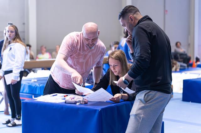 Three coaches intently examine documents at a blue-draped table during an indoor sporting event
