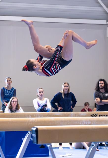 Gymnast performs a backflip mid-air above the balance beam during a competitive routine, spectators watching in the background
