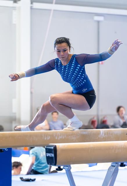 Female gymnast performs a jump on balance beam with arms extended, focused expression, during indoor training session