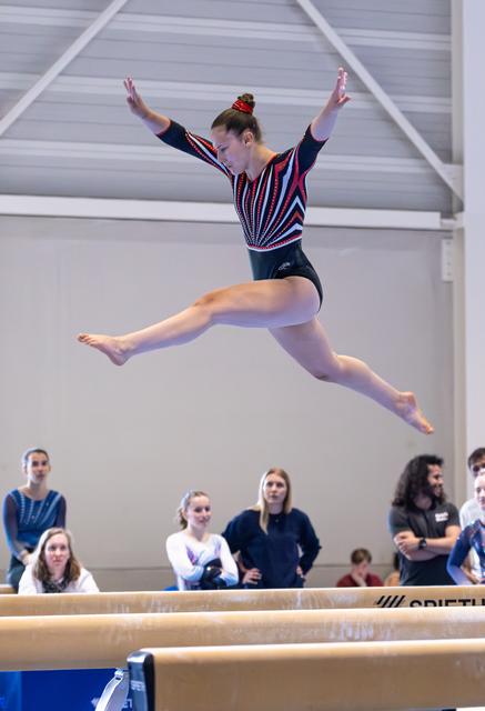 Female gymnast performs a split leap with arms raised above balance beam, spectators watching in background