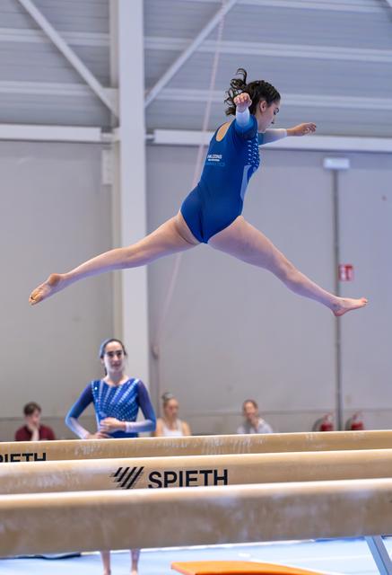 Female gymnast performs dramatic split leap above balance beam with pointed toes during routine at indoor training facility