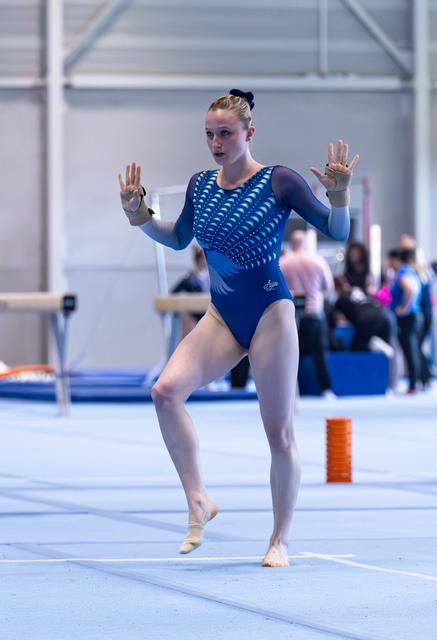 Young athlete performing floor routine in blue sequined leotard with raised hands, showing focused expression during competition