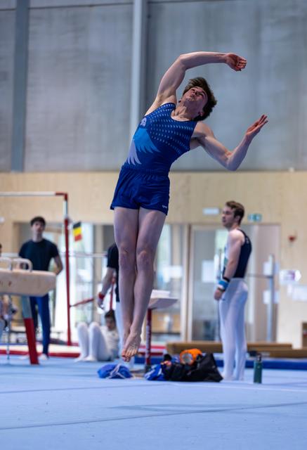Female gymnast performs an elegant arching pose during training, with teammates visible in the background of the gym