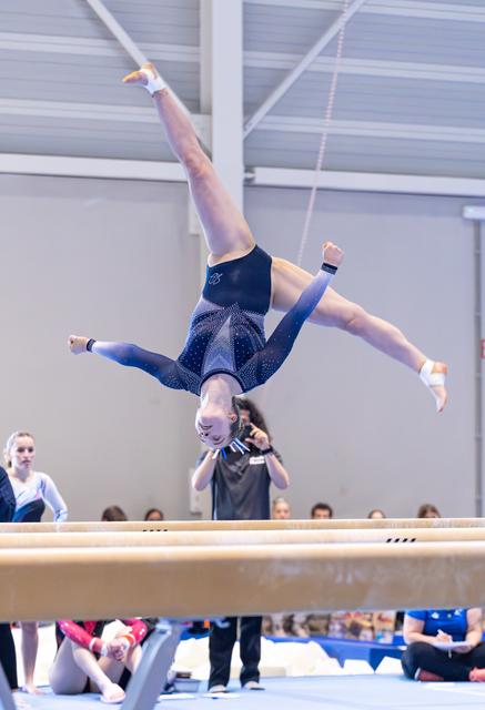Gymnast executes an inverted aerial split above the balance beam while coaches and teammates watch from below