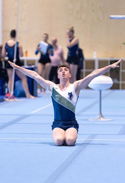 Male gymnast kneeling on floor with arms raised in finishing pose, teammates visible in background during routine