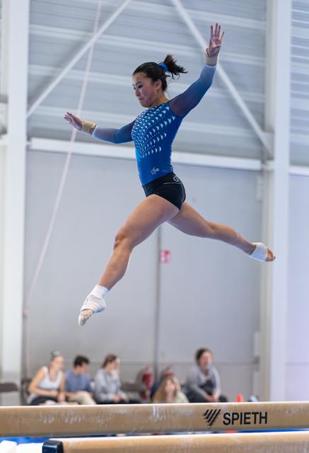 Gymnast performs a split leap high above the balance beam, arms extended gracefully in a blue studded leotard