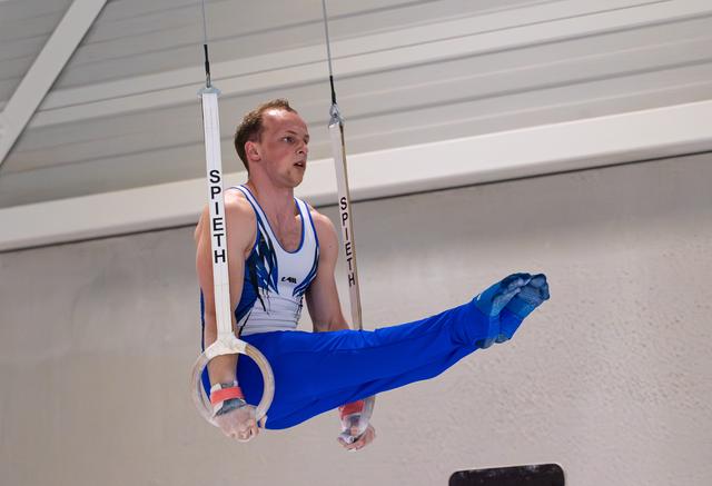 Male gymnast executing a horizontal hold on still rings, legs extended in blue attire, displaying intense concentration