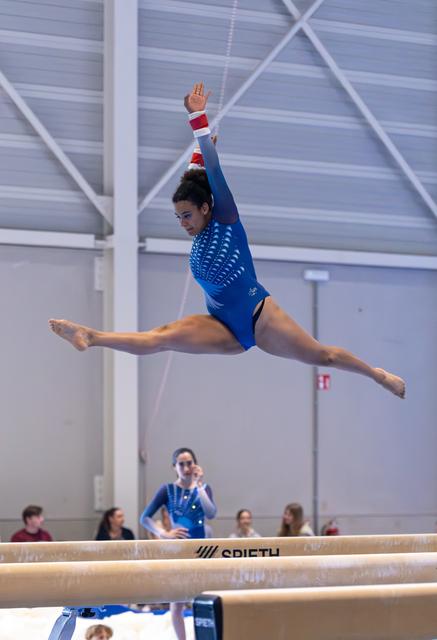 Gymnast executes a split leap with arms extended on balance beam during indoor training, showcasing flexibility and form