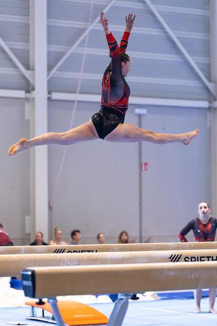 Gymnast performs a perfect split leap over the balance beam with arms raised, showcasing strength and flexibility.