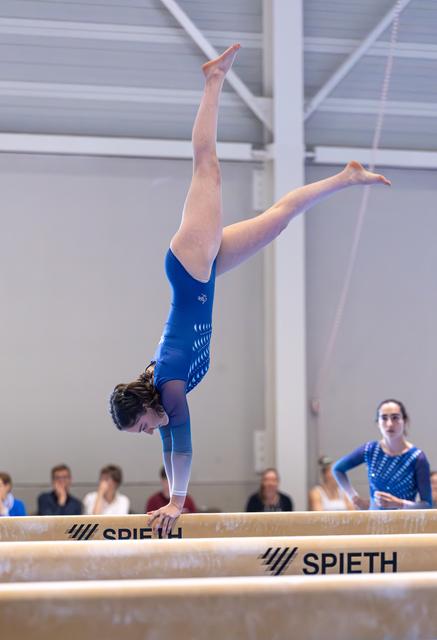 Gymnast performing inverted handstand with vertical leg splits on balance beam during routine at indoor facility