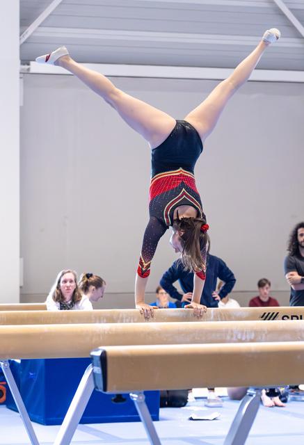 Gymnast performs handstand on balance beam with legs split in V-position during routine at indoor training facility