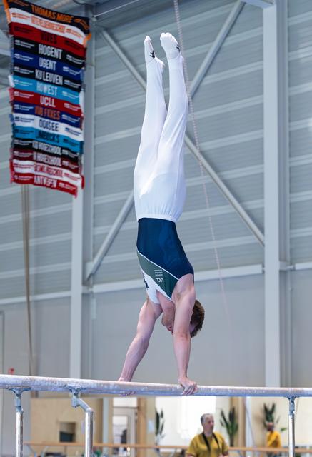 Gymnast performing a handstand on the balance beam with pointed toes, wearing white long-sleeved top and navy shorts