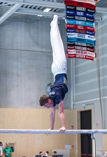 Male athlete performing a handstand on parallel bars in an indoor sports facility with competition signage visible.
