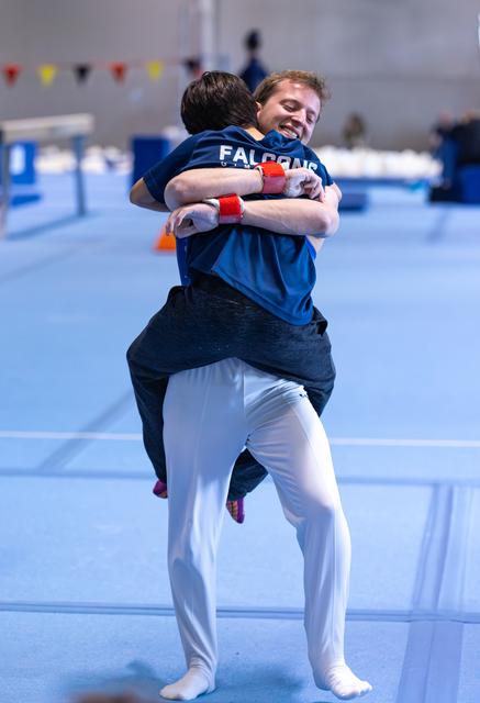 Two teammates share an enthusiastic celebratory hug on the blue training floor, showing pure joy and camaraderie