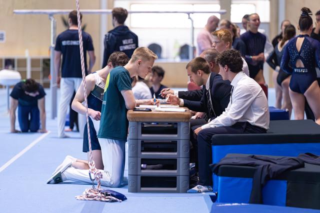 Judges at a table reviewing scores while a young athlete in teal stands beside them, awaiting results during an indoor event