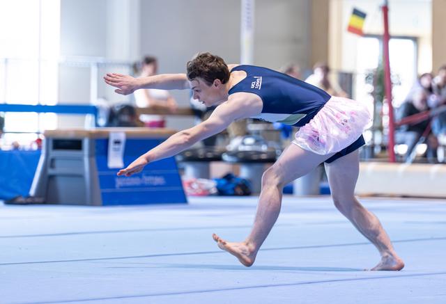 Male athlete performs dynamic leap during floor routine, arms extended with intense concentration at indoor sports facility