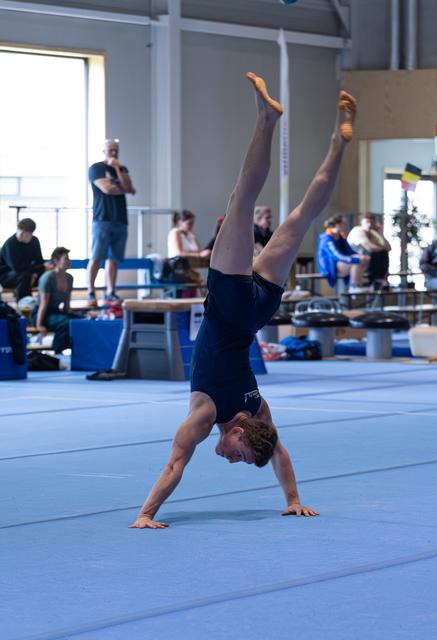 Male athlete performing a handstand with pointed toes on blue mat in training facility with onlookers in background