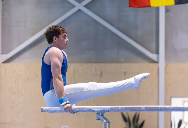Young male athlete performing an L-sit hold on parallel bars, demonstrating strength and control in white uniform and blue leotard